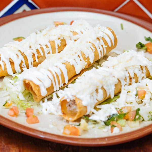 Three golden-brown taquitos topped with white crema, crumbled cheese, and diced tomatoes on a bed of lettuce.