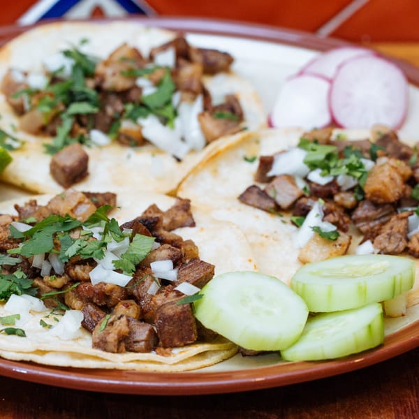 Three Frida Cantina Mexican street tacos with meat, onions, cilantro, and a side of cucumbers and radishes.
