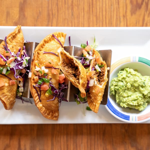 Three golden-brown empanadas, one cut open to show ground beef filling, served with pico de gallo and guacamole.